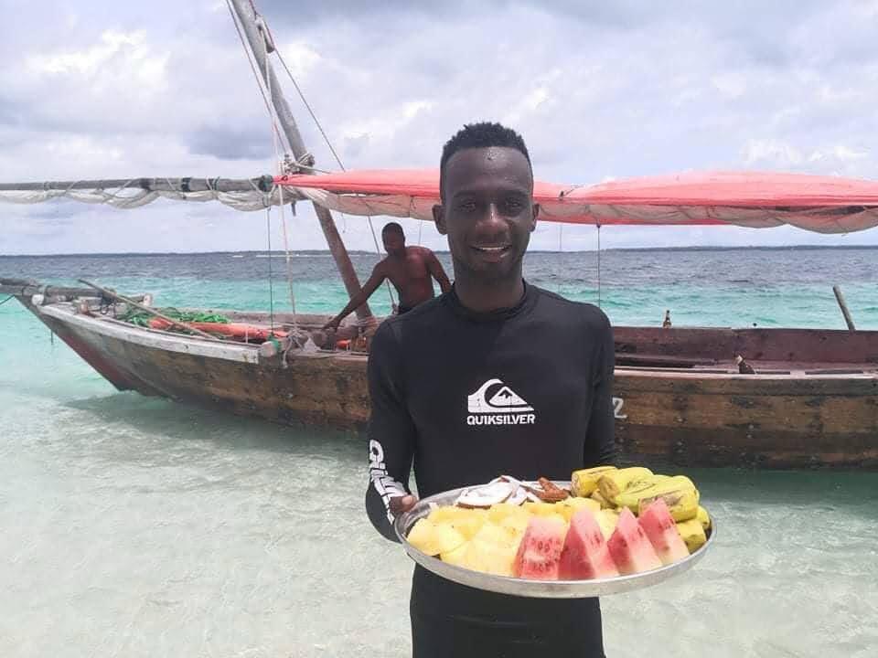 Crew member with a tray of fresh fruit beside a wooden dhow in the lagoon